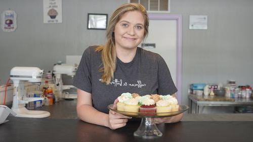 a photograph of Sam Tichnell smiling at the camera, leaning on a countertop with a plate of cupcakes in front of her