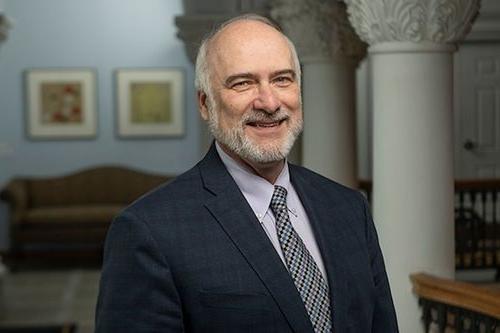 a portrait photograph of Paul Kreider, smiling at the camera in front of a background of white columns and a brown couch